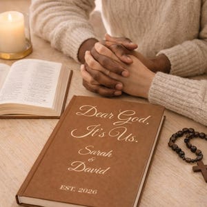 Puede incluir: Un libro marrón con la inscripción "Dear God, It's Us. Sarah & David EST. 2026" se muestra sobre una mesa de madera. Una vela encendida y un libro abierto también están sobre la mesa. Un rosario y dos personas tomadas de la mano están en la imagen.