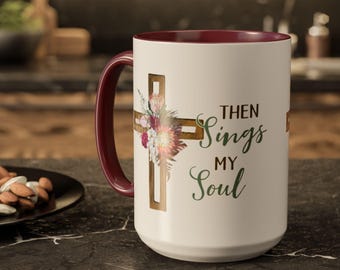 Op de afbeelding: Een witte keramische mok met een donkerrode binnenkant en handvat. De buitenkant is voorzien van een goudkleurige kruisafbeelding versierd met een bloemstuk en de donkergroene tekst "THEN SINGS MY SOUL" in een mix van lettertypes.