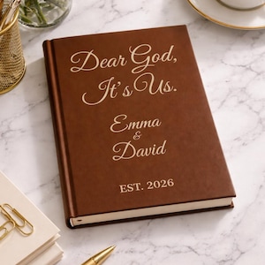 Könnte beinhalten: Ein braunes Buch mit dem Text "Dear God, It's Us." und den Namen "Emma & David" mit "EST. 2026" darunter. Das Buch liegt auf einer Marmoroberfläche, mit einem goldenen Stift, Büroklammern und einer Tasse Kaffee in der Nähe.