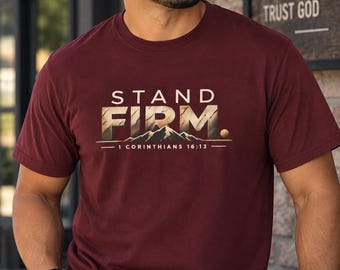 Op de afbeelding: Een bordeauxrood t-shirt met korte mouwen en de tekst STAND FIRM en 1 CORINTHIANS 16:13 boven een bergafbeelding. Het shirt wordt gecombineerd met een bruine en beige baseballpet met mesh-achterkant en de tekst FAITH FAMILY FREEDOM op het voorpaneel.