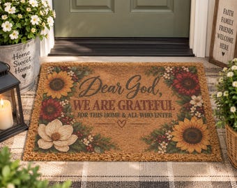 Op de afbeelding: Een rechthoekige deurmat van kokosvezel met een bloemenrand van zonnebloemen, rode bloemen en witte bloesems. De tekst in het midden luidt "Dear God, WE ARE GRATEFUL FOR THIS HOME & ALL WHO ENTER" met een klein hartje onder de tekst.