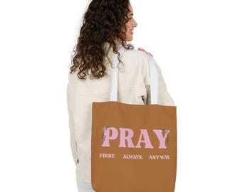 Op de afbeelding: Een bruine tote bag met witte hengsels. De tas toont het woord "PRAY" in roze, met bloemmotieven. De tekst hieronder luidt "FIRST. ALWAYS. ANYWAY."