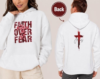 Op de afbeelding: Een witte hoodie met een afbeelding op de voorkant van een kruis gemaakt van roze rozen met de tekst FAITH OVER FEAR in donkerrood. De achterkant van de hoodie toont een rood kruis in aquarel-stijl met het woord Jesus in zwarte letters.