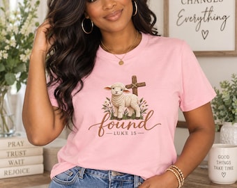 Op de afbeelding: Een lichtroze t-shirt met een afbeelding van een klein lammetje dat voor een houten kruis staat, omringd door wilde bloemen. Onder het lammetje staat het woord found in een bruin cursief lettertype, gevolgd door de tekst LUKE 15.