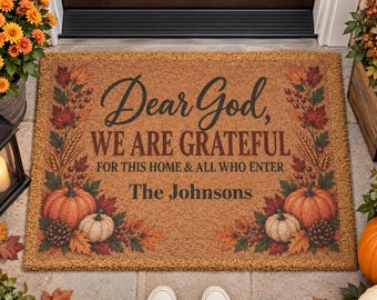 Op de afbeelding: Een rechthoekige beige deurmat van kokosvezel met de zwarte tekst Dear God, WE ARE GRATEFUL FOR THIS HOME & ALL WHO ENTER The Johnsons, omringd door een herfstachtige rand met oranje pompoenen, rode esdoornbladeren en dennenappels.