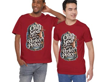 Op de afbeelding: Een donkerrood T-shirt met korte mouwen en een grafische print van biddende handen, een houten kruis en de tekst "Only God can give us Perfect Peace" tegen een achtergrond van bergen en bomen, gecombineerd met een blauwe spijkerbroek.