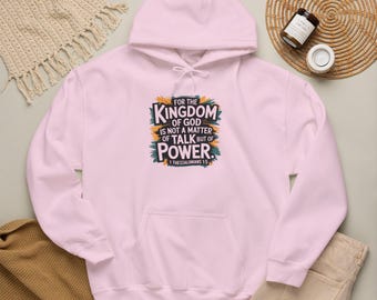 Op de afbeelding: Een lichtroze hoodie met een grafische print op de borst met de tekst: FOR THE KINGDOM OF GOD IS NOT A MATTER OF TALK BUT OF POWER. 1 THESSALONIANS 4:20. De tekst is omringd door kleurrijke, abstracte penseelstreekvormen.