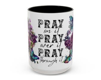 Op de afbeelding: Een witte keramische mok met een zwarte binnenrand, voorzien van een bloemen- en vlinderontwerp in paarse, blauwe en groene tinten. De tekst op de voorzijde luidt: PRAY on it PRAY over it PRAY through it in een combinatie van vetgedrukte en schrijfletters.