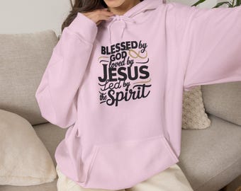 Op de afbeelding: Een lichtroze hoodie met een kangoeroezak aan de voorzijde en een capuchon met trekkoord. Op de borst staat zwarte en gouden tekst met de tekst BLESSED BY GOD LOVED BY JESUS LED BY THE SPIRIT en een afbeelding van een oneindigheidsteken.