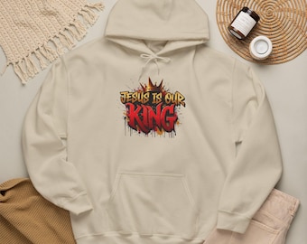 Op de afbeelding: Een beige hoodie met een centrale afbeelding met de tekst "JESUS IS OUR KING" in een gestileerd graffitilettertype. Het ontwerp bevat een kroonillustratie boven de tekst, met rode en oranje verfdruppeleffecten.