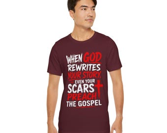 Op de afbeelding: Een bordeauxrood t-shirt met korte mouwen en witte en rode tekst die luidt: WHEN GOD REWRITES YOUR STORY, EVEN YOUR SCARS PREACH THE GOSPEL. Het ontwerp bevat een rood kruissymbool geïntegreerd in de tekst op de voorzijde van het kledingstuk.