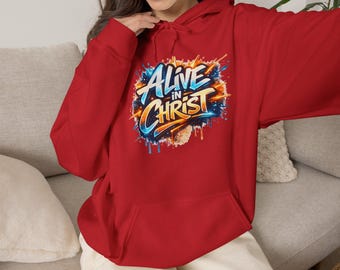Op de afbeelding: Een felrode hoodie met een centrale grafische print met de tekst "ALIVE IN CHRIST" in een gestileerd, graffiti-achtig lettertype. Het ontwerp bevat levendige blauwe, oranje en gele verfspatten en accenten.