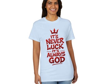 Op de afbeelding: Een lichtblauw t-shirt met korte mouwen en een rode grafische print op de borst. Het ontwerp bevat een kroonicoon boven de tekst: IT'S NEVER LUCK IT'S ALWAYS GOD. De letters hebben een verweerde, getextureerde uitstraling.