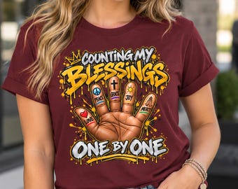 Op de afbeelding: Een bordeauxrood t-shirt met korte mouwen en een afbeelding van een hand met vingers gelabeld Peace, Grace, Joy en Mercy, en een hart gelabeld Love. De tekst luidt Counting My Blessings One By One in gele en witte graffiti-stijl letters.