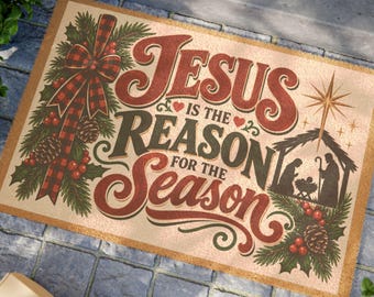 Op de afbeelding: Een rechthoekige beige deurmat met de tekst Jesus is the reason for the season in rode en donkergroene letters. Het ontwerp bevat een rood-zwarte geruite strik, dennentakken, dennenappels, rode bessen en een kerststalafbeelding.