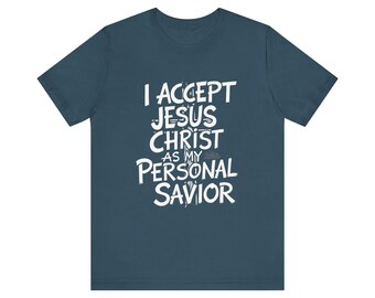 Op de afbeelding: Een donkerblauw t-shirt met korte mouwen en witte tekst op de voorkant met de tekst I ACCEPT JESUS CHRIST AS MY PERSONAL SAVIOR. De letters zijn gestileerd in een vet, handgetekend lettertype en een wit kruis is zichtbaar achter de tekst.