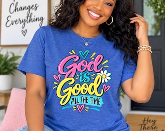 Op de afbeelding: Een blauw T-shirt met korte mouwen en een kleurrijke grafische print met de tekst God is Good ALL THE TIME in roze en gele schrijfletters, geaccentueerd met kleine hartjes, een wit madeliefje en decoratieve gele en blauwe lijnen.
