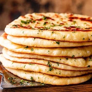 May include: A stack of freshly baked naan bread, golden brown with slightly charred spots, sprinkled with green herbs. The flatbreads are stacked on a wooden cutting board, showcasing a rustic and appetizing presentation.
