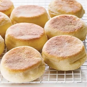 Puede incluir: Primer plano de varios muffins ingleses dorados, recién horneados y colocados sobre una rejilla. Los muffins tienen una parte superior ligeramente texturizada y espolvoreada, con una apariencia suave y esponjosa, ideal para el desayuno.