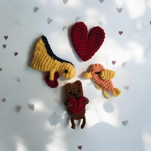 May include: A collection of handmade crochet items, including a red heart, a yellow and blue dinosaur, a brown bear wearing a red sweater, and a peach-colored fish. The items are arranged on a white surface with heart-shaped decorations.