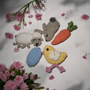May include: A collection of handmade crochet Easter-themed items, including a white sheep, a gray bunny, an orange carrot, a yellow chick with a white flower, and a blue egg. The items are arranged on a white surface with pink flowers.