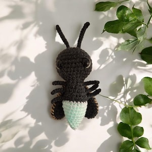 May include: A handmade crochet ant with a black body, large black eyes, and two long antennae. The ant's abdomen is light blue and beige. The ant is displayed on a white surface with green leaves.