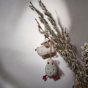 May include: Crocheted Easter ornaments of a sheep and a bird hanging from pussy willow branches. The sheep is white with a tan face, and the bird is gray with red feet. The background is a plain white wall.