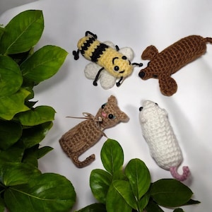 以下が含まれることがあります： 手作りの編みぐるみ動物のおもちゃのコレクション。黄色と黒のミツバチ、茶色のダックスフント、ピンクのリボンが付いた茶色の猫、ピンクの尻尾が付いた白いネズミが含まれています。白い背景です。