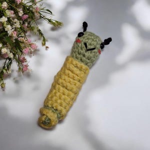 May include: A handcrafted crochet caterpillar toy. The caterpillar features a green head with black antennae and red cheeks. The body is yellow and green. The toy is made of yarn and is designed to resemble a caterpillar.
