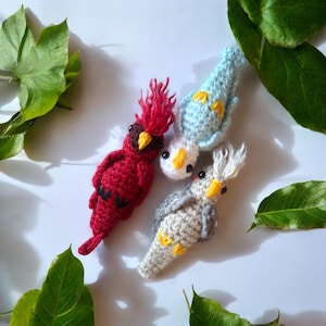 May include: A collection of four handmade crochet birds in various colors. A red cardinal with a spiky crest, a light blue bird with yellow accents, a white bird with yellow accents, and a gray bird with yellow accents. Green leaves are scattered around the birds.