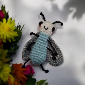 May include: A handcrafted crochet insect with a light blue body, white head, and silver wings. The insect features black antennae, legs, and eyes. The background is white with colourful flowers.
