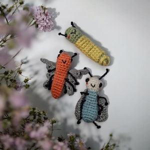 Może przedstawiać: Trzy ręcznie robione zabawki z szydełka: gąsienica, motyl i chrząszcz. Gąsienica jest żółto-zielona, motyl pomarańczowy ze srebrnymi skrzydłami, a chrząszcz niebiesko-biały ze srebrnymi skrzydłami. Zabawki są na białym tle.