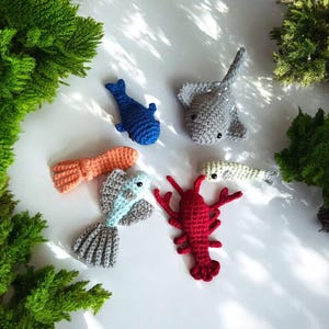 May include: A collection of handmade crochet sea creatures, including a blue whale, gray stingray, orange octopus, red lobster, and a light blue and gray fish. The toys are arranged on a white surface with green foliage in the background.