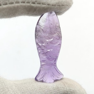 Könnte beinhalten: Eine geschnitzte Amethyst-Fischfigur. Der Fisch hat eine hellviolette Farbe mit detaillierten Schuppen und Flossen. Die Figur wird von einem grauen Stoff gehalten.