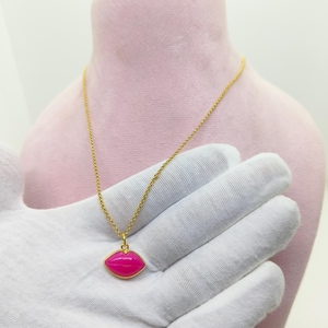 Könnte beinhalten: Eine goldfarbene Halskette mit einem Anhänger in Lippenform, der mit einer leuchtend pinken Emaille-Beschichtung versehen ist. Der Anhänger hängt an einer feinen Gliederkette und zeigt einen lebendigen Kontrast zwischen Gold und Pink.