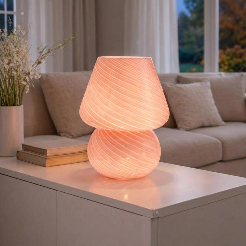 May include: A soft pink glass table lamp with a mushroom-shaped design. The lamp has a round base and a conical shade, both with a swirled pattern. The lamp is lit, emitting a warm glow. The lamp is on a white surface.