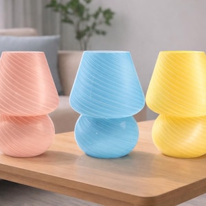 May include: Three decorative mushroom-shaped lamps in pastel colors: pink, blue, and yellow. Each lamp has a rounded base and a conical shade with a swirled pattern. They are arranged on a wooden table.
