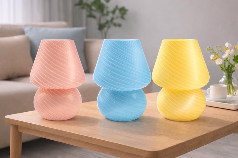May include: Three decorative mushroom-shaped lamps in pastel colors: pink, blue, and yellow. Each lamp has a rounded base and a conical shade with a swirled pattern. They are arranged on a wooden table.