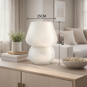 May include: A white, mushroom-shaped table lamp with a swirled glass design. The lamp is 18 cm tall and 15 cm wide. It sits on a light-colored wooden cabinet, next to a small plant and a bowl of decorative objects. Home decor.