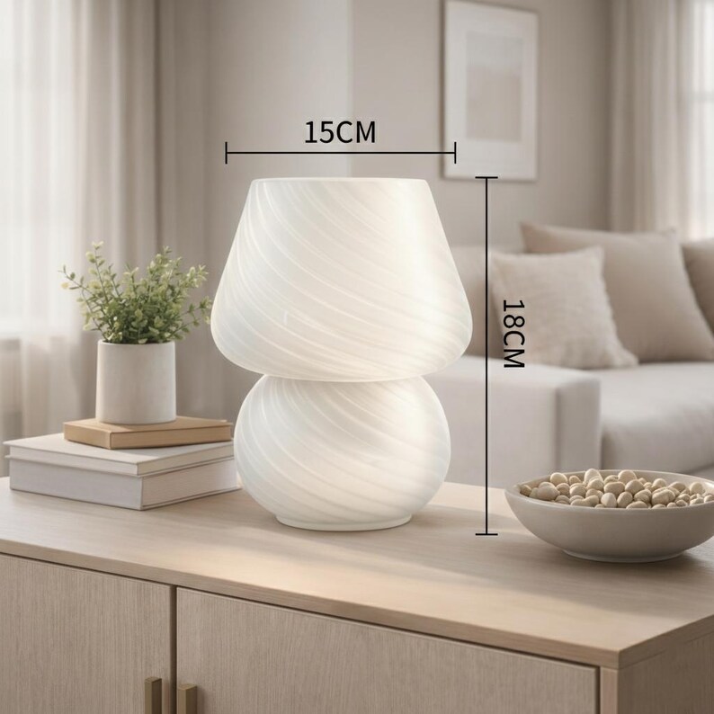 May include: A white, mushroom-shaped table lamp with a swirled glass design. The lamp is 18 cm tall and 15 cm wide. It sits on a light-colored wooden cabinet, next to a small plant and a bowl of decorative objects. Home decor.