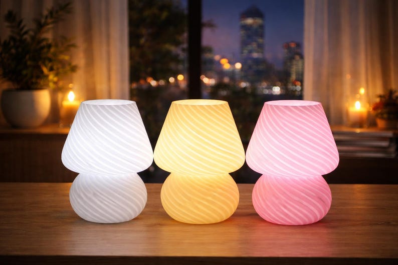 May include: Three mushroom-shaped table lamps in white, yellow, and pink, each with a swirled pattern. The lamps are on a wooden surface, with a cityscape visible through a window in the background.