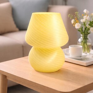 May include: A yellow mushroom-shaped table lamp with a swirled design. The lamp is made of glass and sits on a wooden table. The lamp is approximately 10 inches tall and 7 inches wide. The lamp is a decorative item.