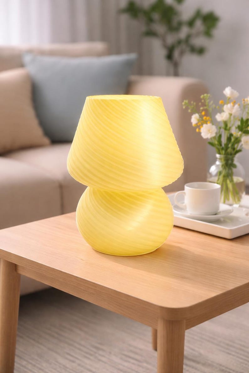 May include: A yellow mushroom-shaped table lamp with a swirled design. The lamp is made of glass and sits on a wooden table. The lamp is approximately 10 inches tall and 7 inches wide. The lamp is a decorative item.