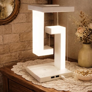 May include: A modern white LED desk lamp with a unique, geometric design. The lamp features a rectangular base with USB ports, a central column, and a floating, illuminated top. The lamp is connected to a power source via a white cable.