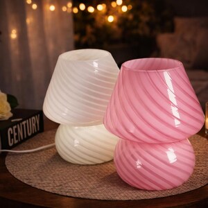 May include: Two glass mushroom-shaped lamps, one white and one pink, sit on a round table. The lamps have a swirled design. A lit candle in a glass holder and a book are also on the table. The background is softly lit.
