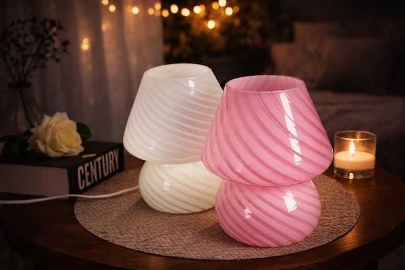 May include: Two glass mushroom-shaped lamps, one white and one pink, sit on a round table. The lamps have a swirled design. A lit candle in a glass holder and a book are also on the table. The background is softly lit.