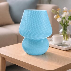 May include: A light blue glass mushroom-shaped lamp with a textured, swirled design. The lamp has a rounded base and a flared top, sitting on a wooden table. The lamp is a decorative lighting fixture.