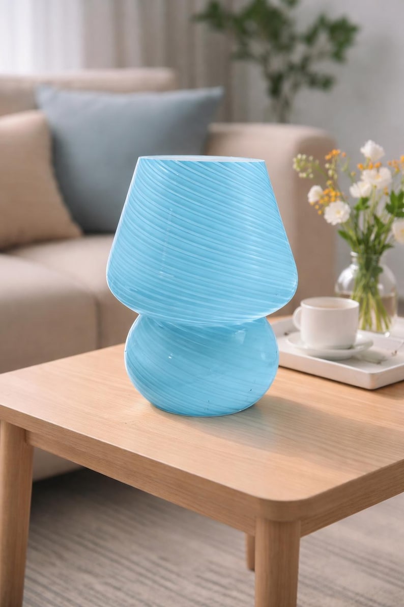 May include: A light blue glass mushroom-shaped lamp with a textured, swirled design. The lamp has a rounded base and a flared top, sitting on a wooden table. The lamp is a decorative lighting fixture.