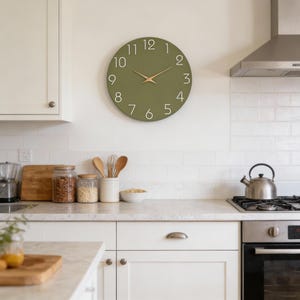 Può includere: Un orologio da parete rotondo verde oliva con numeri bianchi e lancette dorate. L'orologio è montato su una parete bianca sopra un piano di lavoro della cucina. La cucina ha mobili bianchi e una cappa aspirante in acciaio inossidabile.