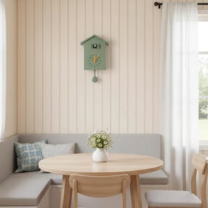 May include: A sage green cuckoo clock with a pendulum hangs on a cream-colored wall. The clock has a house-shaped design with a round face and black numerals. The clock is part of a dining room setting with a round wooden table and a gray bench.